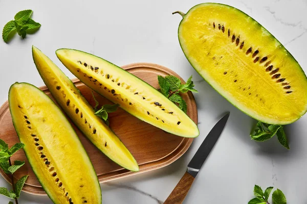 Top view of cut delicious exotic yellow watermelon with seeds on wooden chopping board with knife and mint on marble surface — Stock Photo