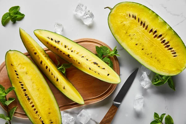 Top view of cut delicious exotic yellow watermelon with seeds on wooden chopping board with knife, mint and ice cubes on marble surface — Stock Photo