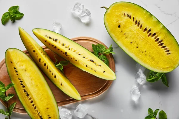 Draufsicht auf geschnittene köstliche exotische gelbe Wassermelone mit Samen auf Marmoroberfläche mit Minze, Eis und Holzhackbrett — Stockfoto