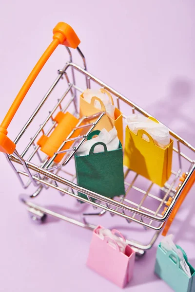 Carrito de compras decorativo con bolsas de papel de colores sobre fondo violeta - foto de stock