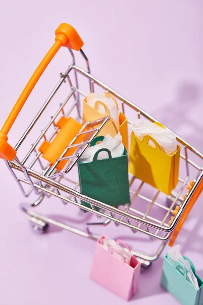 Decorative shopping cart with colorful paper bags on violet background — Stock Photo