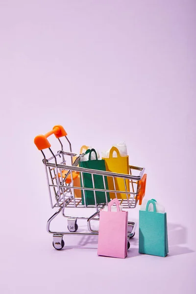 Toy cart with colorful paper shopping bags on violet background — Stock Photo