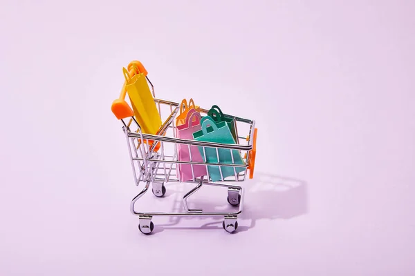 Toy shopping cart with colorful paper bags on violet background — Stock Photo