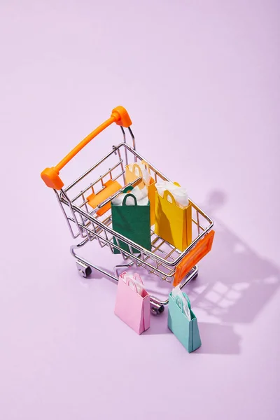Algunas pequeñas bolsas de papel cerca del carro de juguete con coloridas bolsas de compras sobre fondo violeta — Stock Photo