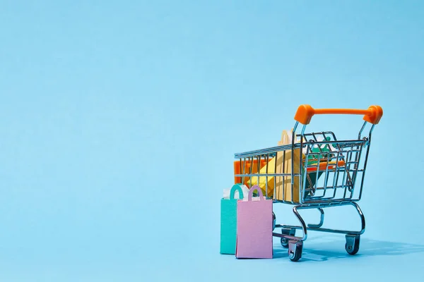 Quelques petits sacs en papier près du chariot de jouets avec des sacs à provisions colorés sur bleu — Photo de stock