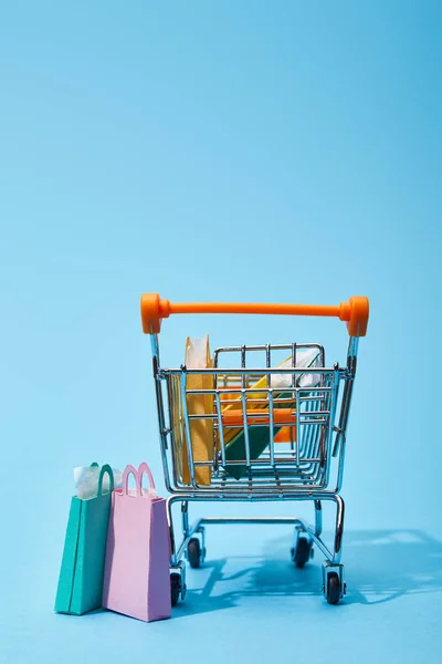 Algunas pequeñas bolsas de papel cerca del carro de juguete con coloridas bolsas de compras sobre fondo azul - foto de stock