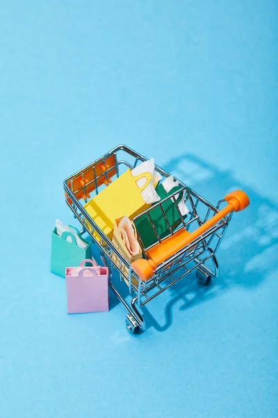 Chariot à jouets avec des sacs en papier colorés près de quelques sacs à provisions sur fond bleu — Photo de stock