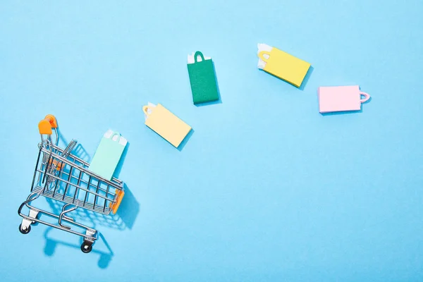 Vista superior del carrito de compras de juguetes cerca de coloridas bolsas de papel en azul - foto de stock