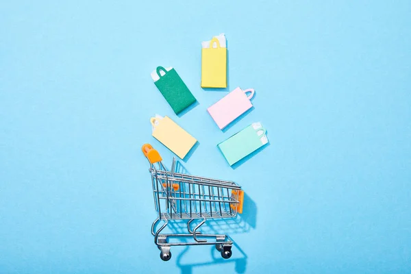 Top view of colorful paper bags falling into toy shopping cart on blue — Stock Photo