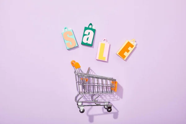 Vista superior de coloridas bolsas de juguete con letras de venta cerca del carrito de compras en violeta - foto de stock