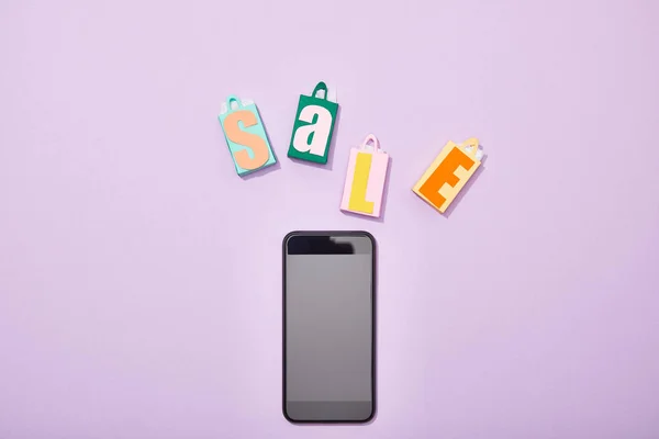 Вид сверху на разноцветные бумажные пакеты с надписью рядом со смартфоном с пустым экраном на фиолетовом — стоковое фото