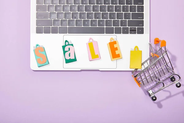 Top view of colorful shopping bags with sale lettering on laptop near small toy cart on violet — Stock Photo