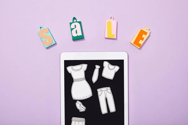 Vista superior de sacos de compras de papel com venda lettering perto de corte de papel de roupas em tablet digital em violeta — Fotografia de Stock