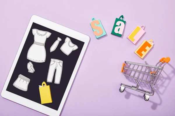 Vista superior de corte de papel de roupas em tablet digital perto de sacos de compras com lettering venda caindo no carrinho de brinquedo em violeta — Fotografia de Stock