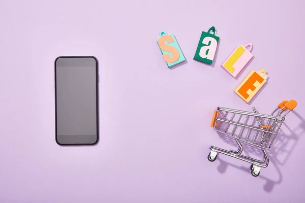 Vista superior do smartphone perto de sacos de compras com letras de venda caindo no carrinho de brinquedo em violeta — Fotografia de Stock