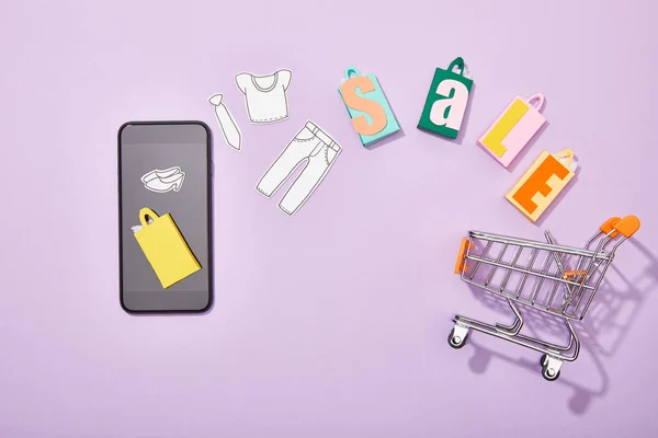 Vista superior do smartphone perto de sacos de compras com lettering venda e corte de papel de roupas caindo no carrinho de brinquedo em violeta — Fotografia de Stock
