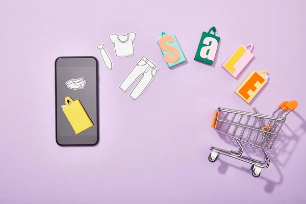 Вид смартфона рядом с пакетами для покупок с надписью и разрезанной бумагой, падающей в игрушечную корзину на фиолетовой — стоковое фото