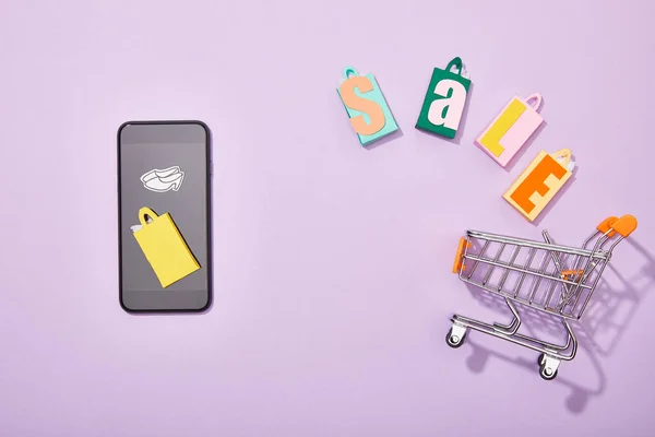 Vista superior del teléfono inteligente cerca de las bolsas de compras con letras de venta cayendo en el carro de juguete en violeta, concepto de e-shopping - foto de stock