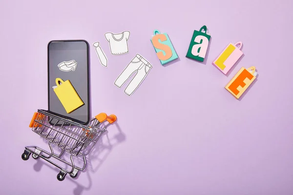 Вид зверху смартфона з паперовим вирізом одягу та жовтим паперовим мішком, що падає в іграшковий кошик біля барвистих торговельних мішків з продажем на фіолетовому — стокове фото