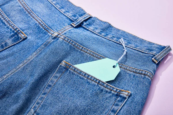 Закрыть вид бирюзовой бумажной бирки на джинсах на фиолетовом — стоковое фото
