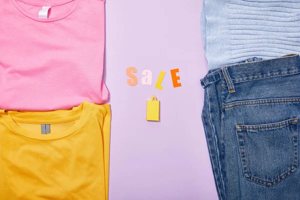 Top view of sale lettering above shopping paper bag near colorful clothes on violet — Stock Photo