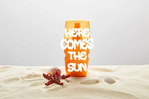 Protector solar en botella de color naranja sobre arena con estrellas de mar sobre fondo gris con aquí viene la inscripción del sol - foto de stock