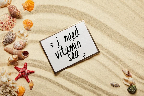 Vista superior do cartão com eu preciso de vitamina mar lettering perto conchas, estrelas do mar vermelho e corais na praia arenosa — Fotografia de Stock
