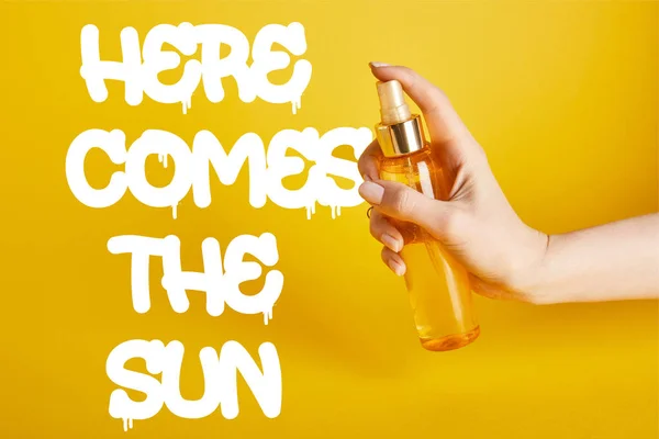 Vista recortada de la mujer sosteniendo la botella con el aerosol del protector solar en el fondo amarillo con aquí viene la letra del sol - foto de stock