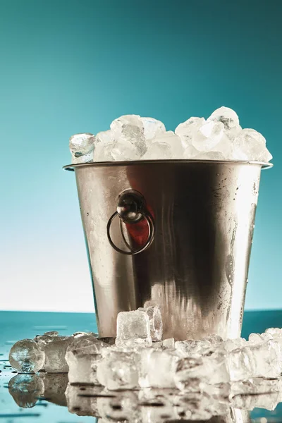 Transparent ice cubes in metal bucket near water puddles on emerald and white background — Stock Photo