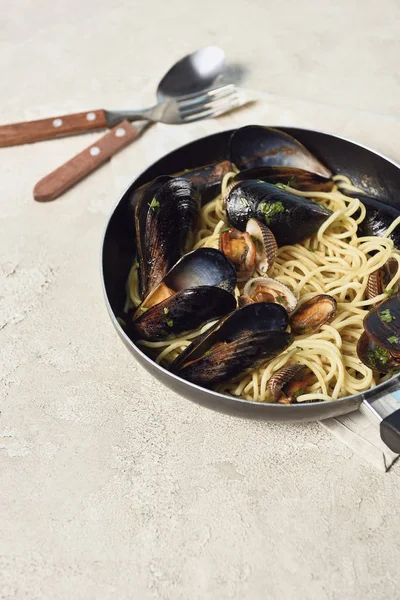 Köstliche Pasta mit Meeresfrüchten in der Pfanne in der Nähe von Besteck auf strukturiertem grauen Hintergrund — Stockfoto