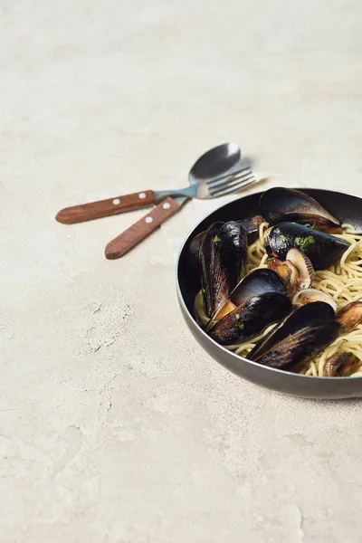Leckere Pasta mit Meeresfrüchten in der Pfanne in der Nähe von Besteck auf strukturiertem grauen Hintergrund — Stockfoto
