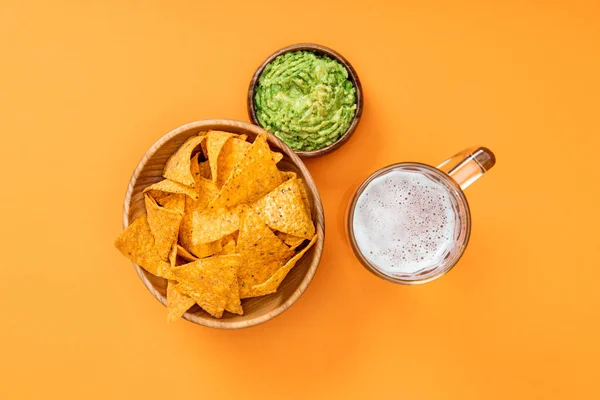 Vista dall'alto di nachos croccanti in ciotola di legno vicino guacamole e birra su sfondo arancione, cucina messicana — Foto stock