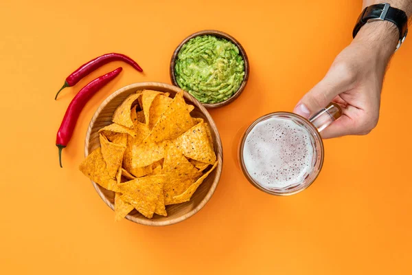 Vista cortada do homem segurando vidro de cerveja perto de nachos crocantes e guacamole no fundo laranja, cozinha mexicana — Fotografia de Stock