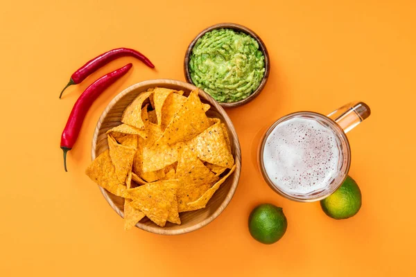 Draufsicht auf knusprige Nachos, Guacamole, Bier, Limetten und Chilischoten auf orangefarbenem Hintergrund, mexikanische Küche — Stockfoto