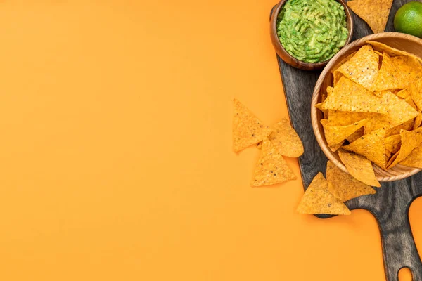 Top view of crispy Mexican nachos on wooden cutting board, guacamole and lime on orange background with copy space — Stock Photo