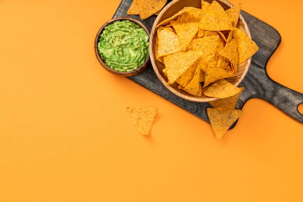 Top view of crispy Mexican nachos served with guacamole on wooden cutting board on orange background — Stock Photo