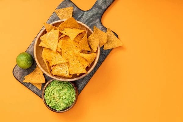 Top view of crispy Mexican nachos served with guacamole and lime on wooden cutting board on orange background — Stock Photo