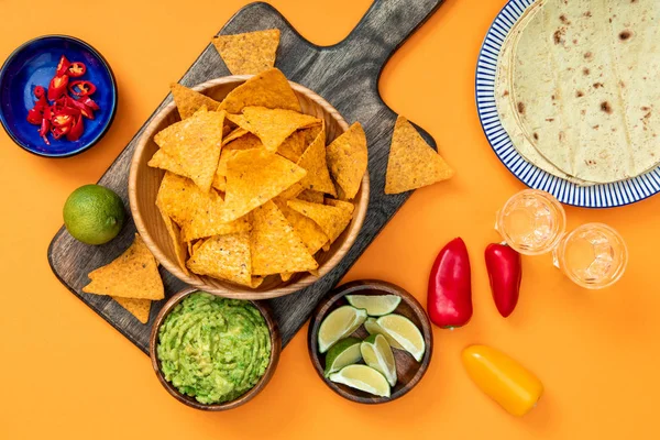 Top view of crispy Mexican nachos served on wooden cutting board with guacamole, peppers, limes, Tequila and tortillas on orange background — Stock Photo