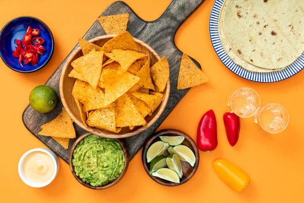 Nachos mexicanos servidos em tábua de corte de madeira com guacamole, molho de queijo, pimentas, limas, tequila e tortilhas em fundo laranja — Fotografia de Stock