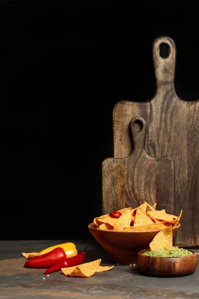 Nachos mexicanos con sal, guacamole y chiles cerca de tablas de cortar de madera en mesa de piedra aislada en negro - foto de stock