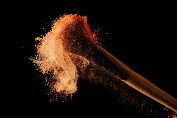 Cosmetic brush with orange powder explosion on black background — Stock Photo