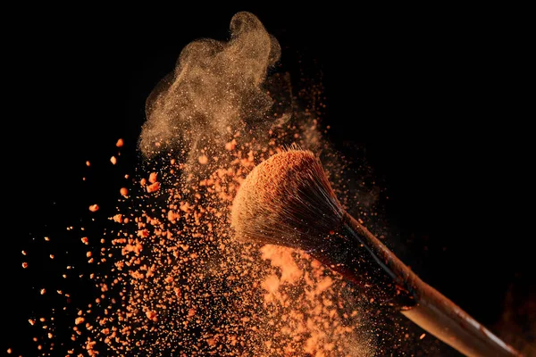 Cosmetic brush with bright orange powder explosion on black background — Stock Photo