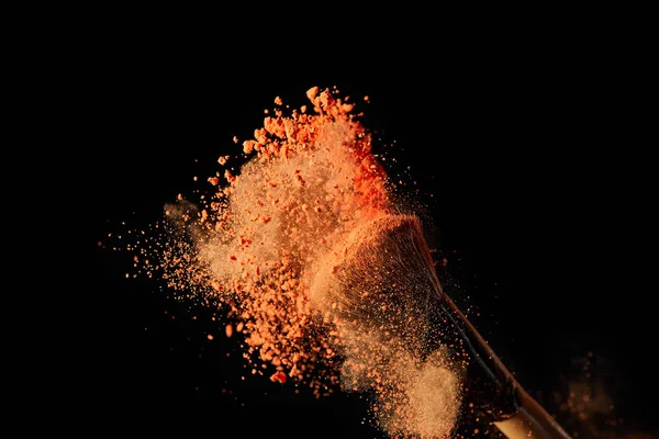 Cosmetic brush with colorful orange powder explosion on black background — Stock Photo