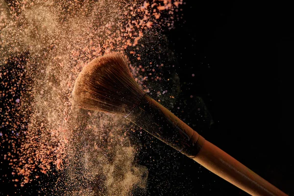 Soft cosmetic brush with colorful orange powder explosion on black background — Stock Photo