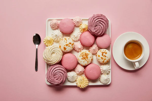 Poser à plat avec des meringues et des macarons assortis sur un plat carré avec une tasse de café et une cuillère sur fond rose — Photo de stock