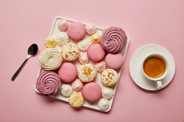 Vue de dessus des meringues et des macarons sur plat carré avec cuillère et tasse de thé sur fond rose — Photo de stock