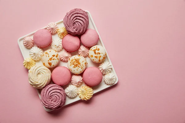 Vista superior de macarrones franceses surtidos y merengues en plato cuadrado sobre fondo rosa - foto de stock