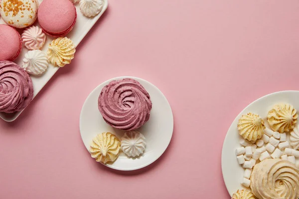 Soft pink zephyr with small meringues on saucer with macaroons and marshmallows on plates on pink background — Stock Photo