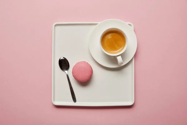 Top view of pink macaroon with spoon and cup of coffee on big square white dish on pink background — Stock Photo
