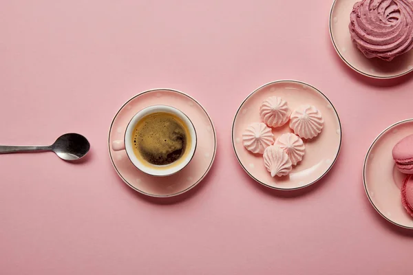 Top view of pink meringues and macaroons on pink dotted saucers with spoon and cup of coffee on pink background — Stock Photo